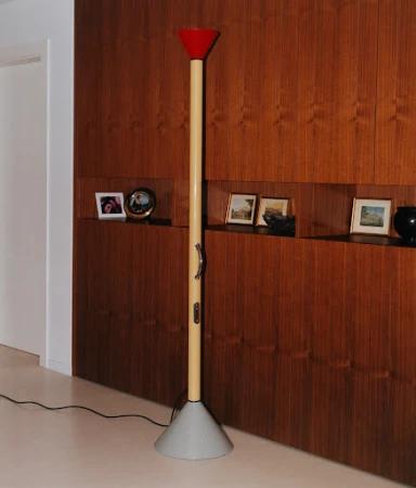 Tall floor lamp with red top and grey base stands against a wood-paneled wall with inset shelves displaying photos and decorative objects.