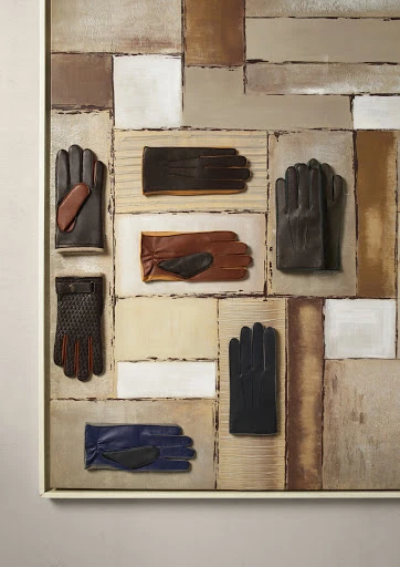 Still life painting featuring multiple pairs of leather gloves in shades of brown, black, and blue, arranged on a patchwork background.