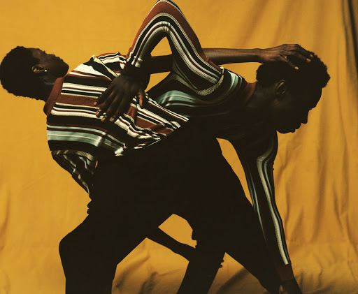 Two black men with striped shirts pose against a yellow backdrop in a studio fashion shot, showcasing artistic expression and style.
