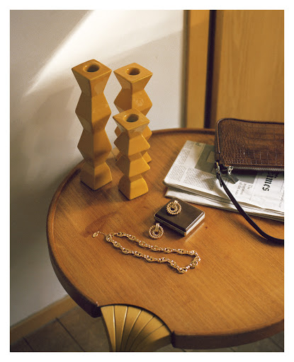 Still life of wooden candle holders, gold jewelry, a clutch purse, and a newspaper on a round wooden table indoors with natural lighting.