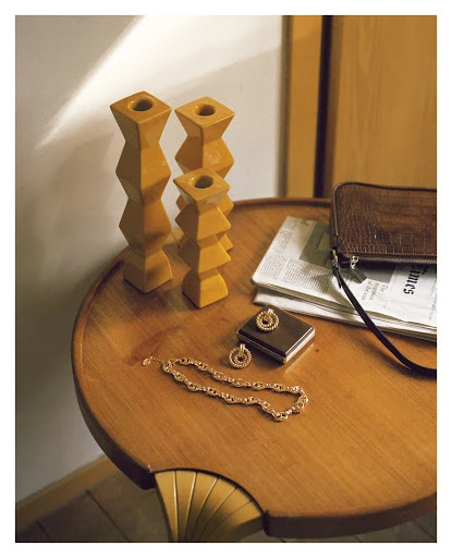 Still life of wooden candle holders, gold jewelry, a clutch purse, and a newspaper on a round wooden table indoors with natural lighting.