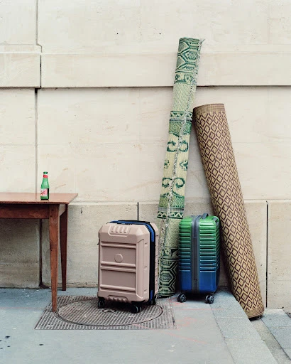 Two rolling suitcases and two rolled-up rugs are positioned against a textured wall outside, next to a small wooden table and drink bottle.