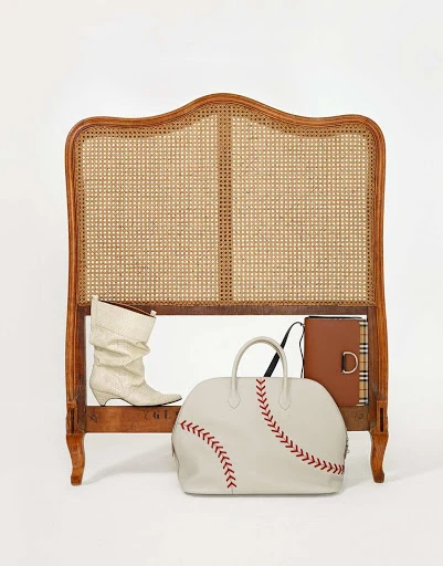 Fashion still life featuring a tan wicker headboard showcasing a white leather boot, baseball-inspired bag, and leather portfolio.