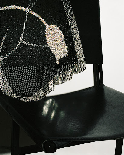 Close-up of a black leather chair draped with a black mesh fabric featuring silver glitter designs, against a neutral backdrop.