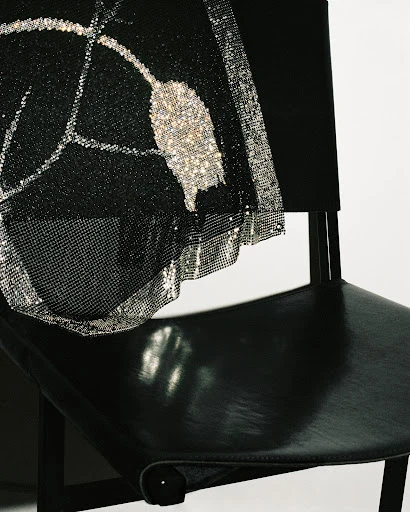 Close-up of a black leather chair draped with a black mesh fabric featuring silver glitter designs, against a neutral backdrop.