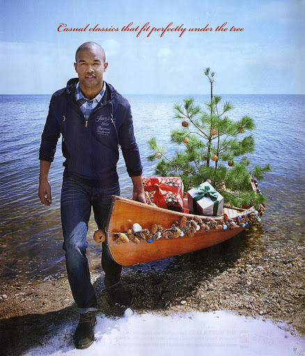 Adult male walks on a beach near water carrying a canoe filled with presents and a Christmas tree, creating a festive holiday scene.