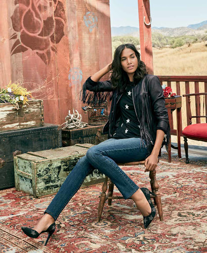 Woman sits on a chair in a star patterned top, leather jacket, and jeans in front of a rustic backdrop with countryside views.