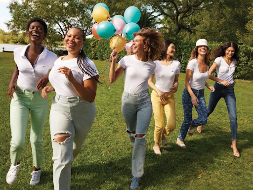 Group of six multi-ethnic women in jeans and white tops running on grass, smiling, and holding balloons in an outdoor setting.