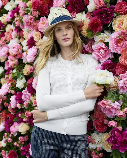 A young woman wearing a white cardigan and hat poses against a wall of pink and red flowers, holding white flowers in her hand.