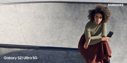 A stylish adult female model with curly hair posing in a skatepark, holding a smartphone, wearing a turtleneck and long skirt.