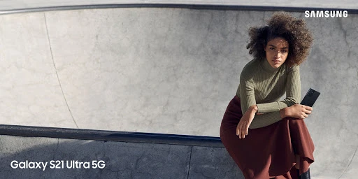 A stylish adult female model with curly hair posing in a skatepark, holding a smartphone, wearing a turtleneck and long skirt.
