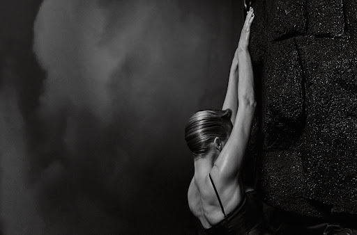 Monochrome image featuring an adult female athlete in active pose, climbing dark rock formation under dramatic lighting against stormy backdrop.