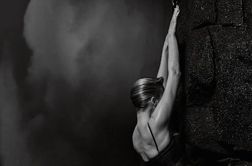 Monochrome image featuring an adult female athlete in active pose, climbing dark rock formation under dramatic lighting against stormy backdrop.