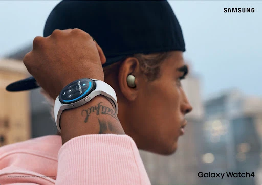 Close-up of a person wearing a Samsung Galaxy Watch4 with white wristband and wireless earbuds, set against a blurred cityscape backdrop.