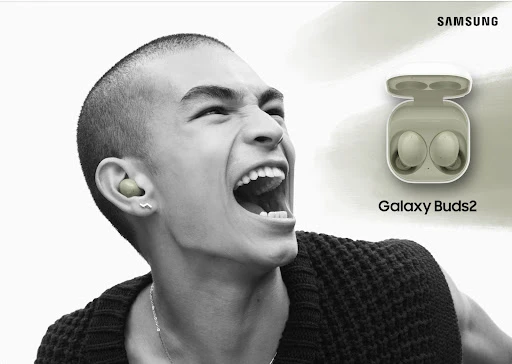 A person with short hair is seen wearing wireless earbuds, next to a pair of wireless earbuds in their charging case with Samsung logo.