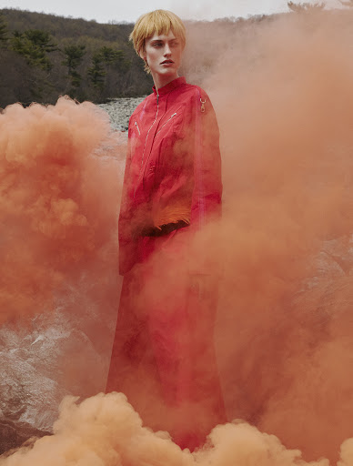 Person in a red outfit surrounded by orange smoke in an outdoor setting, with trees and natural rocks visible in the background.