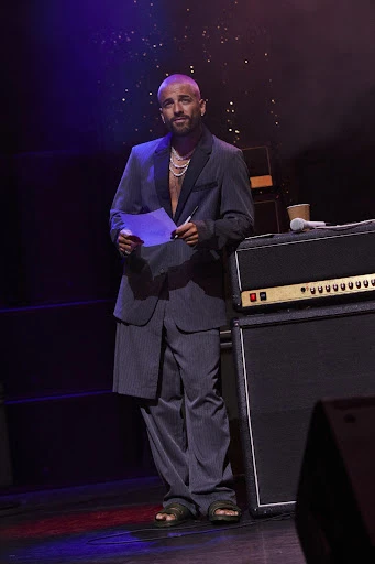 Bald man in a grey pinstripe suit standing on stage with papers and a microphone next to a speaker at an event. 
