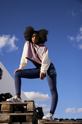 Woman with afro wearing Adidas hoodie and leggings poses on wooden pallets with blue sky and white building in the background.