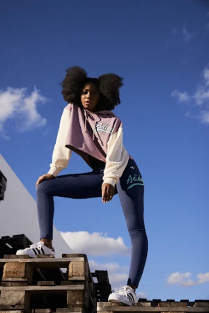 Woman with afro wearing Adidas hoodie and leggings poses on wooden pallets with blue sky and white building in the background.