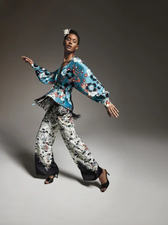 A woman in a stylish patterned outfit poses in motion, showcasing a modern take on fashion with a floral blouse and wide-leg pants.