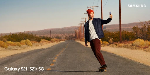 Young man on skateboard on desert road, taking selfie with Samsung Galaxy S21 phone, showcasing mobile technology and outdoor lifestyle.