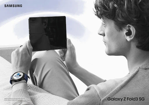 Man holding a black Samsung Galaxy Z Fold3 5G phone, wearing wireless earbuds and a smartwatch in a grey-scale promotional studio shot.