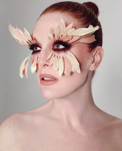 Close-up portrait of a woman with fair skin and red hair styled in a bun, wearing elaborate feather eyelashes and dramatic makeup.