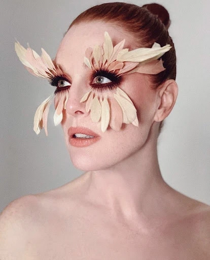 Close-up portrait of a woman with fair skin and red hair styled in a bun, wearing elaborate feather eyelashes and dramatic makeup.