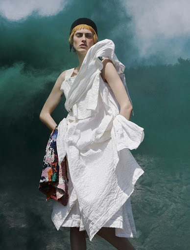 Woman in a white dress with an expressive pose against an abstract turquoise background with light cloud-like formations, fashion photography.
