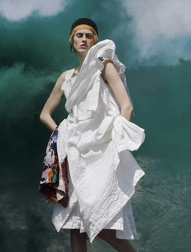 Woman in a white dress with an expressive pose against an abstract turquoise background with light cloud-like formations, fashion photography.