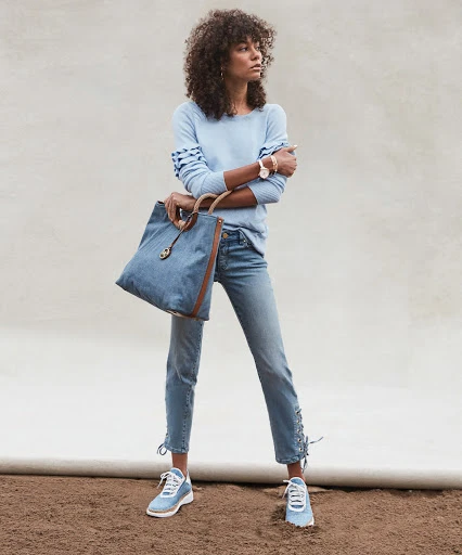 Full-length shot of an adult female model wearing a light blue sweater, jeans, and sneakers, holding a blue tote bag in a studio setting.