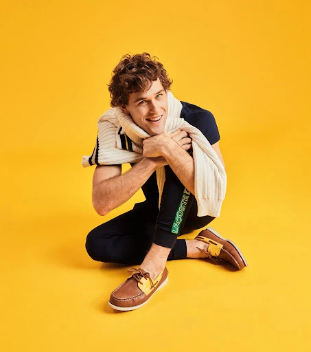 A stylish male model poses against a vibrant yellow background, showcasing fashion apparel and boat shoes. The casual yet fashionable studio shot.