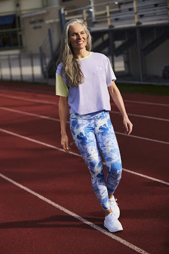 Adult woman with long grey hair poses in sportswear on a red running track with white lines during daylight. Sport and fitness concept.