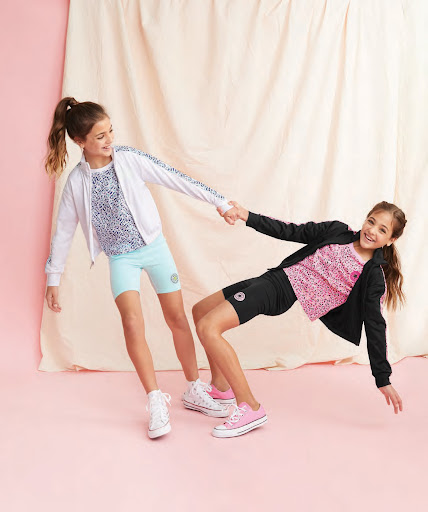 Two young girls pose playfully in colorful athletic wear against a draped backdrop, showcasing a lively, energetic fashion scene.