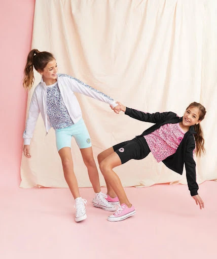 Two young girls pose playfully in colorful athletic wear against a draped backdrop, showcasing a lively, energetic fashion scene.
