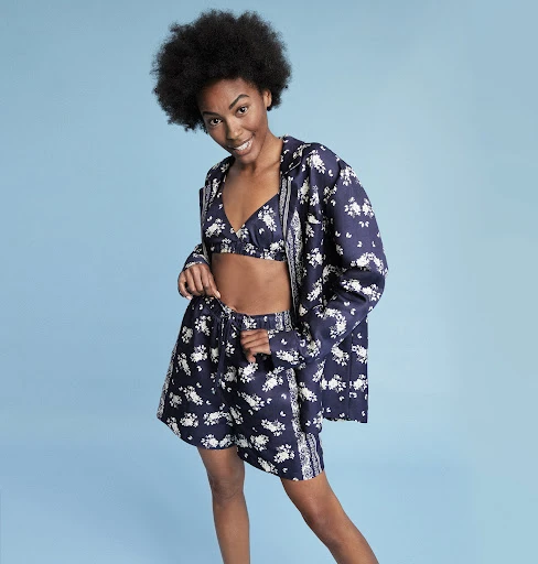 Smiling African-American woman in a patterned navy blue bikini top, shorts, and hooded jacket on a light blue background in a studio.