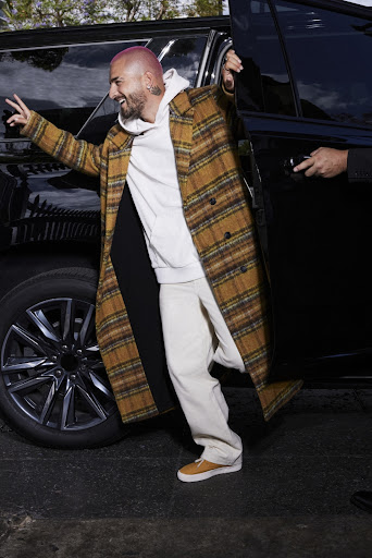 Man exiting a black SUV, wearing a plaid coat over a white hoodie, khaki pants, and sneakers, with a playful expression and pink hair.