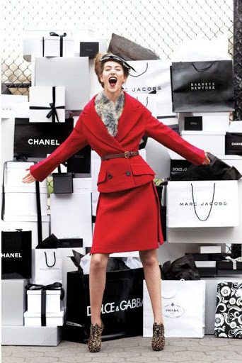 Fashion model poses in red suit surrounded by designer shopping bags in an outdoor setting, expressing joy and excitement.