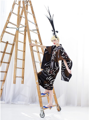A woman in a kimono-style dress and feathered headpiece poses on a wooden ladder in a studio, creating a high-fashion editorial shot.