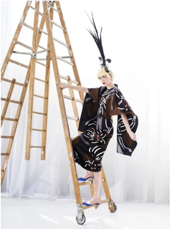 A woman in a kimono-style dress and feathered headpiece poses on a wooden ladder in a studio, creating a high-fashion editorial shot.