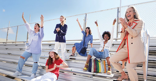 Enthusiastic group of adult women of various ethnicities, laughing and cheering from stadium bleachers on a sunny day.