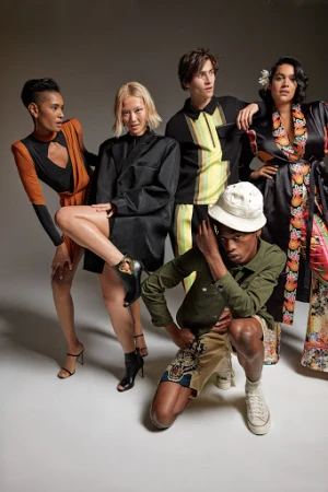 Group of five fashion models in a photo studio wearing various outfits, including dresses, blouses, shorts, and heels against a grey background.