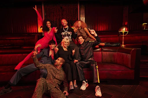 Group of seven people posing on a burgundy leather couch in a dimly lit room with red accents, showcasing fashion and entertainment.
