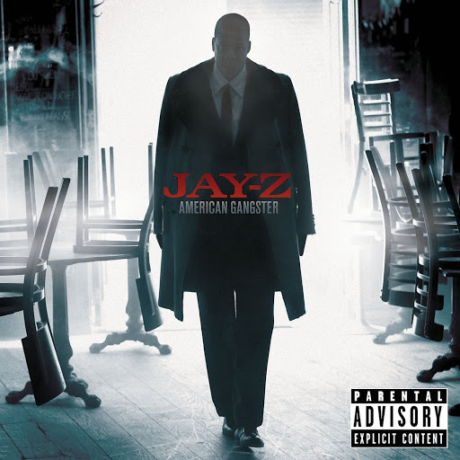Jay-Z stands in a stark, cinematic album cover image featuring a dimly lit interior with overturned tables, dressed in a dark suit and coat.