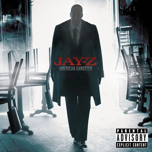 Jay-Z stands in a stark, cinematic album cover image featuring a dimly lit interior with overturned tables, dressed in a dark suit and coat.