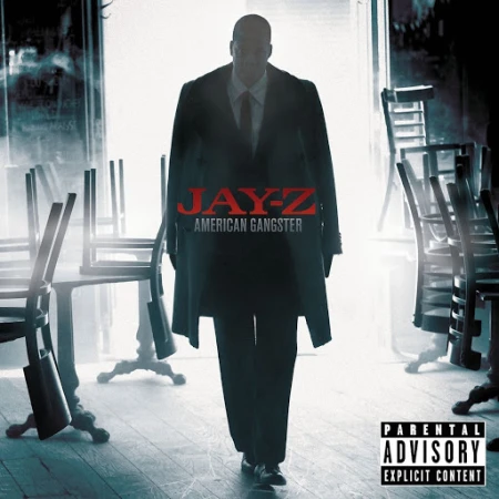 Jay-Z stands in a stark, cinematic album cover image featuring a dimly lit interior with overturned tables, dressed in a dark suit and coat.