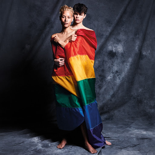 Two models wrapped in a colorful rainbow flag posing in a photo studio with dark background, portrait studio shot.