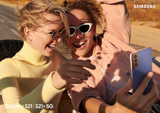 Two cheerful adults, fair-skinned, wearing sunglasses and casual tops, smile and hold a blue Samsung Galaxy S21 smartphone in a candid outdoor shot.