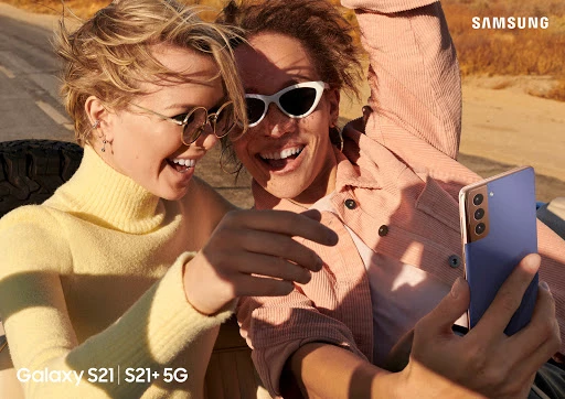 Two cheerful adults, fair-skinned, wearing sunglasses and casual tops, smile and hold a blue Samsung Galaxy S21 smartphone in a candid outdoor shot.