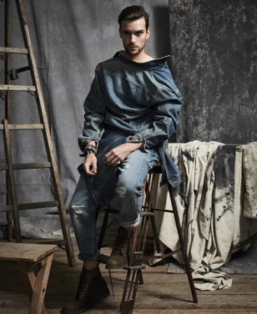 Man wearing a denim jacket and jeans sits on a wooden stool in a photo studio with a ladder and canvas in the background.